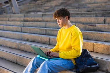 Genç adam basamaklarda otururken dizüstü bilgisayar kullanıyor, çevrimiçi öğrenimine odaklanıyor, temiz havanın ve şehir ortamının tadını çıkarıyor.
