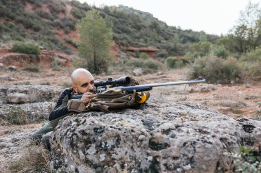 Avcı yere yatar, tüfeğiyle nişan alır doğal bir ortamda sırt çantasında dinlenir.