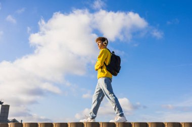 Sırt çantası ve kulaklıkları olan genç bir erkek öğrenci, parlak mavi gökyüzünün altında, tüylü beyaz bulutlarla birlikte bir duvarda yürüyor.