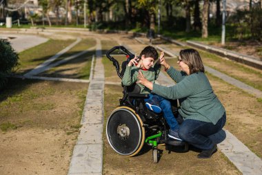 Anne, beyin felci yüzünden tekerlekli sandalye kullanan oğluyla şakalaşıyor. Parkta güneşli bir günün tadını çıkarıyor.