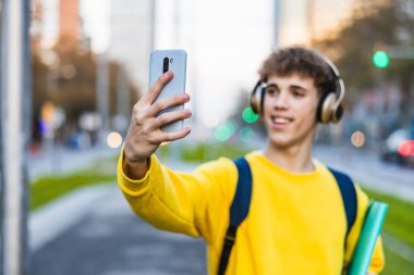 Kulaklık takan ve kitap taşıyan mutlu bir üniversite öğrencisi şehirde akıllı telefonuyla selfie çekiyor.