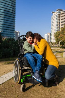 Anne engelli oğluna sarılıyor tekerlekli sandalyede oturuyor, şehirde güneşli bir günün tadını çıkarıyor.