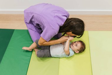 Optometrist kadın, yeşil bir paspasın üzerinde yatan bir çocuğun görme yetisini incelemek için retinoskop kullanıyor.