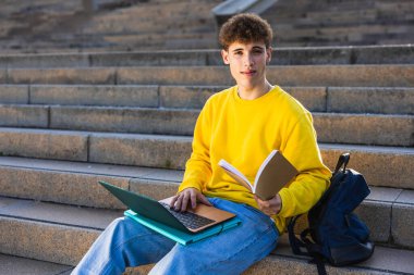 Genç adam basamaklarda otururken dizüstü bilgisayar kullanıyor ve kitap okuyor, teknoloji ve geleneksel öğrenimi birleştiriyor.