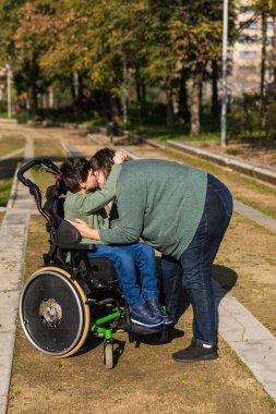 Anne sakat bir şekilde oğlunu öpüyor. Tekerlekli sandalyede oturuyor. Sevgisini, ilgisini ve desteğini ifade ediyor.