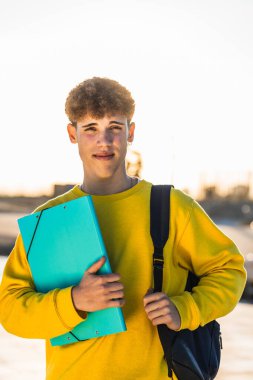 Sırt çantası ve dosya taşıyan genç bir üniversite öğrencisi kampüste verimli bir günün ardından güneşin sıcacık günbatımının tadını çıkarıyor.