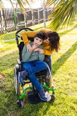 Sevgi dolu anne, güneşli bir parkta tekerlekli sandalyede oturan dokuz yaşındaki engelli oğlunu kucaklıyor.