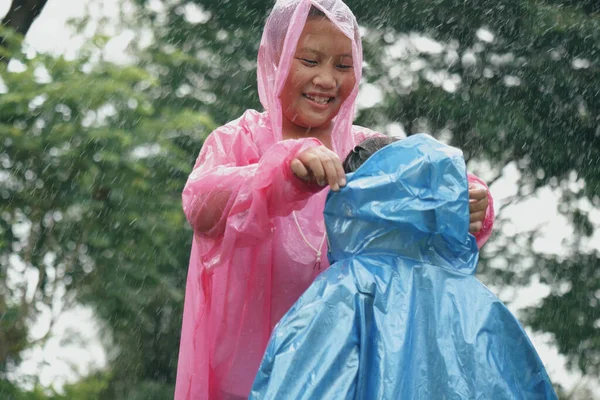 Cheerful Asian boy with beautiful smile in pink raincoat helps his little sister wearing head cover raincoat in rain surrounded by nature and trees. A kind-hearted child is the future of sustainability thinking people.