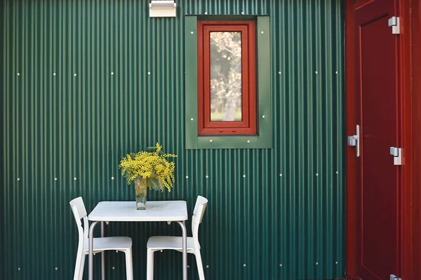Green and red exterior of a container house with furnished entrance and flowers.