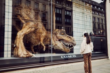 Londra, İngiltere - 25 Mart 2025: Bir kadın Londra şehir merkezindeki Koç mağazasının vitrininde dev bir dinozor heykeli görüyor.