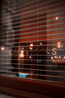 Londra, İngiltere - 28 Eylül 2024: Retro Cocktails tabelası Venedik jaluzilerinde sıcak iç ışıklar ve şık bir Londra bar ortamında karamsar yansımalarla görülüyor