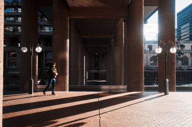 Londra, İngiltere - 28 Eylül 2024: Bir kişi Barbican Malikanesi 'nin sütunlu geçidinde dramatik gölgeler ve tarihi St. Giles Kilisesi ile su üzerinde görünür bir şekilde yürüyor.