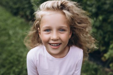  Sarı saçlı ve mavi gözlü küçük kız bahçede mutlu bir şekilde oynuyor, doğanın ve temiz havanın tadını çıkarıyor. Çocukların sağlığı ve neşesi. Mutlu çocukluk