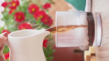 Sabahları kırsal bir ortamda Amerikan kahvesi. Kadın cam bardağa bir sürahi ile espresso kahvesi dolduruyor. Açık havanın tadını çıkarıyorum. Dikey video 4k, yaşam tarzı.