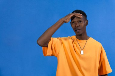 Meraklı bir adam, uzaklığa bakarken gözleri koruyor, canlı mavi arkaplanda bir şey arıyor ya da arıyor.