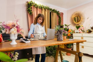Çiçekçi dizüstü bilgisayar kullanırken not alıyor, çiçekçi dükkanında online siparişleri yönetiyor, renkli buketler ve bitkilerle çevrili.