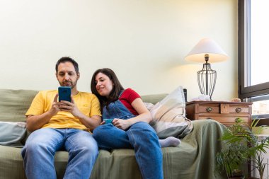 Kafkasyalı çift kanepede birlikte rahatlıyor. Sosyal medyada akıllı bir telefonla geziniyorlar ve bir fincan kahvenin tadını çıkarıyorlar.