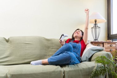 Mutlu genç bir kadın kanepede uzanıyor ve evinde video oyunu oynarken kolunu kaldırıyor, zaferi kutluyor.