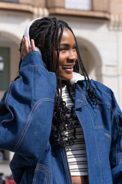 Şık genç bir kadın kulaklıkla müzik dinliyor, gülümsüyor ve bir caddede dikilirken yüzünü çeviriyor.