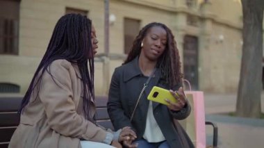 İki neşeli Afrikalı kadın bir bankta otururken akıllı telefonuyla selfie çekiyor.