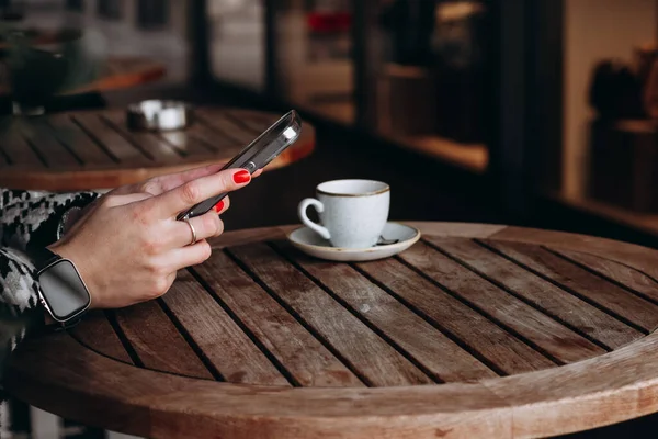 stock image Women's hands with red manicures holding a cell phone at a table in a cafe with a cup of coffee