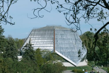 Çatı ve duvarlarında üçgen cam paneller olan büyük, açısal bir sera ağaçlarla çevrili bir şehir parkında yer almaktadır.