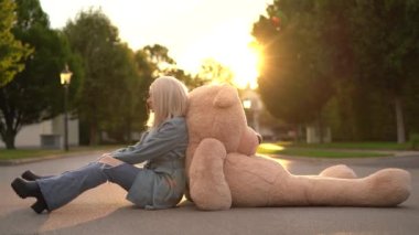 Blonde girl in blue wear sit in the street with big bear toy, sunset time, beautiful view. High quality 4k footage