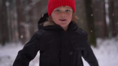 Kırmızı şapkalı küçük çocuk zıplıyor ve kameraya bakıyor, kar ormanı, kış sezonu, 4k video. Yüksek kalite 4k görüntü