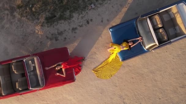 車のフードに横たわる2人の女の子の空中映像 赤いドレスを着た女の子が黄色のドレスを着ている 女の子は車の上に横たわる カッパドキアの日没時間 — ストック動画