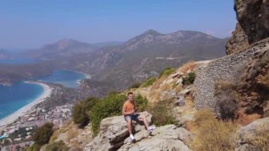 Genç adamın panoramik görüntüsü kayanın üzerinde uzaktan kumandalı drone ile oturup kendini havaya ateş ediyor. Oludeniz plajı geçmişi. - Evet. Yüksek kalite 4k görüntü