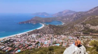 Genç adam uzaktan kumandalı drone ile kayanın üzerinde oturuyor, ayarlıyor, kendini havaya ateş ettiriyor. Oludeniz plajı geçmişi. İnsansız hava aracından inanılmaz güzel panoramik manzara. Yüksek kalite 4k görüntü