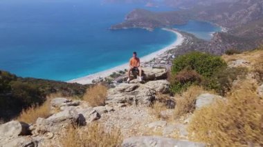 Young man sit on the rock with remote control drone , shooting himself aerial. Oludeniz beach background. Amazing beautiful panoramic view from drone of natural park. High quality 4k footage
