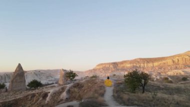 Gün batımında Kapadokya dağlarında kuyruğu olan uzun sarı bir elbiseyle dolaşan uzun boylu bir kız modelin güzel drone görüntüleri. Tam HD görüntü. Gün batımı görünümü