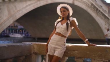 Şapkalı ve beyaz elbiseli bir kız nehir kanalı boyunca kalır, İtalya, Venedik kanalı, gün batımı saatinde. Yüksek kalite 4k görüntü