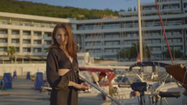 Siyah elbiseli, uzun saçlı mutlu bir kadın limanda bir teknenin yanında oturuyor. Kış manzarasının tadını çıkarırken gülümsüyor. Arka planda bir şehir silüeti var.