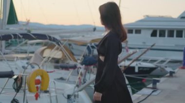 Siyah elbiseli bir kadın limandaki bir teknenin yanında durup gülümsüyor. Su birikintisinin tadını çıkarırken gökyüzü su gemisinden yansıyor.
