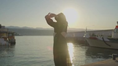 Gün batımında, bir kadın iskelede suyun kenarında duruyor. Sahne, gökyüzünün, suyun ve güneş ışığının güzelliğini yakalar. Huzurlu ve huzurlu bir atmosfer yaratır.