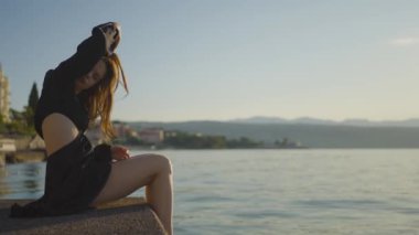 Siyah saçlı bir kadın göle bakan bir iskelede mutlu bir şekilde oturuyor, huzurlu manzaranın ve gökyüzünün tadını çıkarıyor. Bu doğal ortamda rahat ve mutlu görünüyor.
