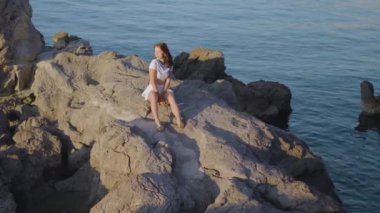 Beyaz elbiseli bir kadın, güneşli bir günde kayalık bir uçurumda oturmuş, denizin huzurlu manzarasının tadını çıkarıyor.