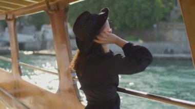 Siyah şapkalı şık bir kadın, bir tekne güvertesinden manzaranın tadını çıkarıyor, huzurlu anı yakalıyor..
