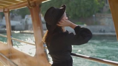 Siyah şapkalı şık bir kadın, bir tekne güvertesinden manzaranın tadını çıkarıyor, huzurlu anı yakalıyor..