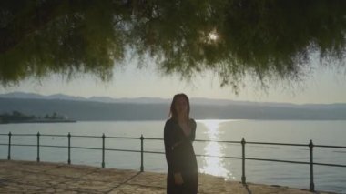 Huzurlu bir gün batımı manzarası, gölün yanında bir kadının silueti, bir ağacın altında duruyor, dingin bir hava..