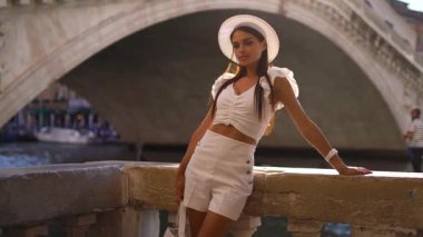 Şapkalı ve beyaz elbiseli bir kız nehir kanalı boyunca kalır, İtalya, Venedik kanalı, gün batımı saatinde. Yüksek kalite 4k görüntü