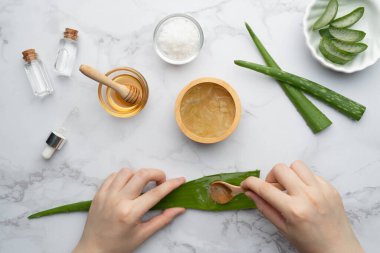 Natural homemade aloe vera gel face and body scrub for skin problems. DIY Recipe for sunburn, acne and dry skin. Top view of aloe vera, honey, sea salt and essential oil bottle on marble background.