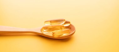 Sarı arka planda ahşap kaşıkta yağlı kapsüller. Besin takviyesi balık yağı, omega 3, omega 6, omega 9, e vitamini, d vitamini ve a vitamini içerir..