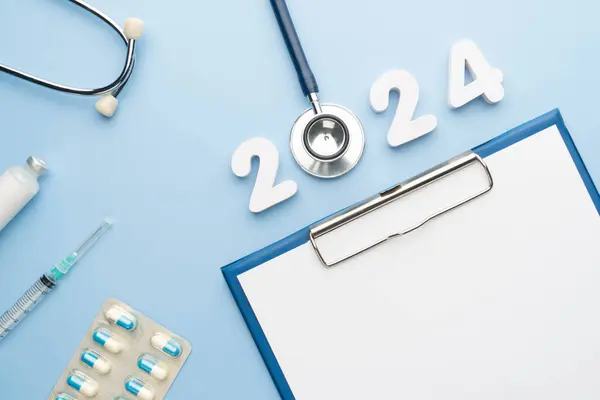 2024 Happy New Year banner for health care and medical concept. Stethoscope with doctor order chart, prescription, pills, syringe, vaccine vial and white number 2024 on table blue background.