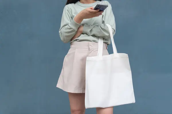 Blank white tote bag canvas fabric with handle mock up design. Close up of woman holding eco or reusable shopping bag and using smartphone near blue metalic wall. No plastic bag and ecology concept.