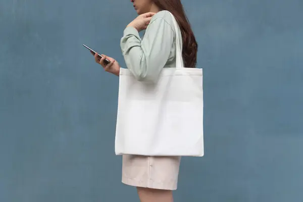 Blank white tote bag canvas fabric with handle mock up design. Close up of woman holding eco or reusable shopping bag and using smartphone near blue metalic wall. No plastic bag and ecology concept.