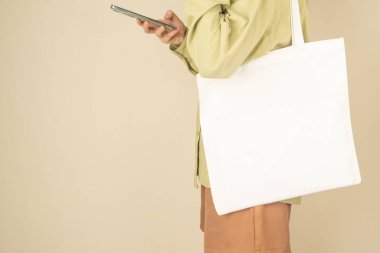 Boş beyaz çanta bez kumaş ve kabzası da tasarım. Çevre dostu ya da tekrar kullanılabilir bir alışveriş çantası taşıyan ve krem rengi duvarın yanında akıllı telefon kullanan kadınlara yakın durun. Plastik poşet ve ekoloji konsepti yok.
