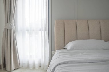 Perdelerin arasından süzülen yumuşak güneş ışığıyla yatak odası sahnesi. Modern minimalizm tarzı yatak odası içi bej ve gri tonlarda..